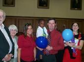 A group of people, including a couple holding balloons, gather in a courthouse setting, celebrating the adoption of two children.