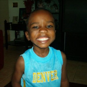 A young boy with a big smile wearing a blue tank top with 'DENVER' printed on it, standing indoors.