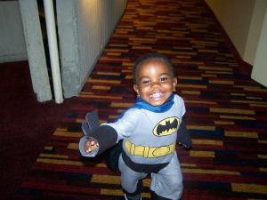 A young boy dressed as Batman, smiling and running down a colorful carpeted hallway.