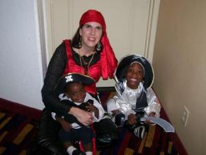 A woman dressed as a pirate sits on the floor next to two young boys, also dressed in pirate costumes, in a hotel hallway.