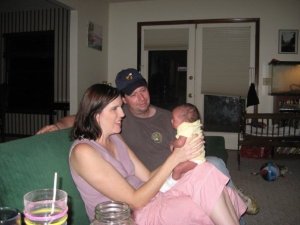 A mother and father sitting on a couch, gazing at a newborn baby being held by the mother, with a cozy living room setting in the background.