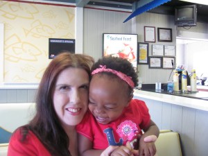 A woman with long hair smiling while holding a young girl with curly hair, both wearing red shirts. They are in a brightly lit interior of a restaurant, with a menu visible in the background.