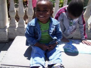 A smiling young child sitting on a stone surface, wearing a denim jacket and green shirt, with another child partially visible in the background examining a map.