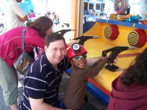 A man and a young boy wearing a helmet engage in a fun game at an amusement park, smiling and interacting with each other.