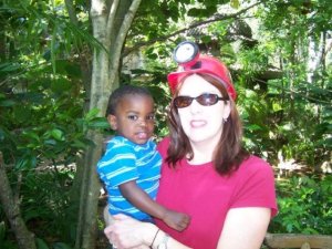 A woman wearing sunglasses and a red shirt is holding a young child in a blue striped shirt. They are surrounded by lush greenery, suggesting a natural outdoor setting.