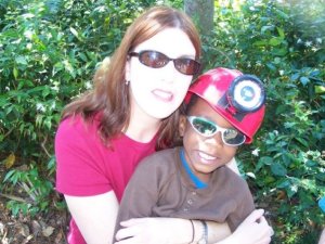 A woman with sunglasses and a red shirt hugs a young boy in a red hat and sunglasses, both smiling against a green leafy background.