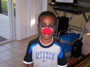 A young boy wearing oversized glasses and a red clown nose, smiling while inside a room with light-colored walls and tiled flooring.