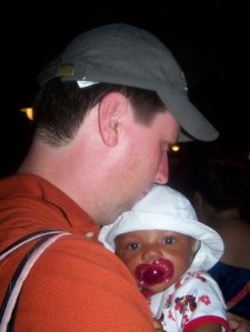 A man wearing a cap holds a young child with a pacifier, both facing towards the camera.