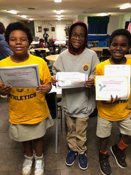 Three children proudly holding their award certificates in a school setting. Two are wearing bright yellow shirts, while one is in a gray hoodie.
