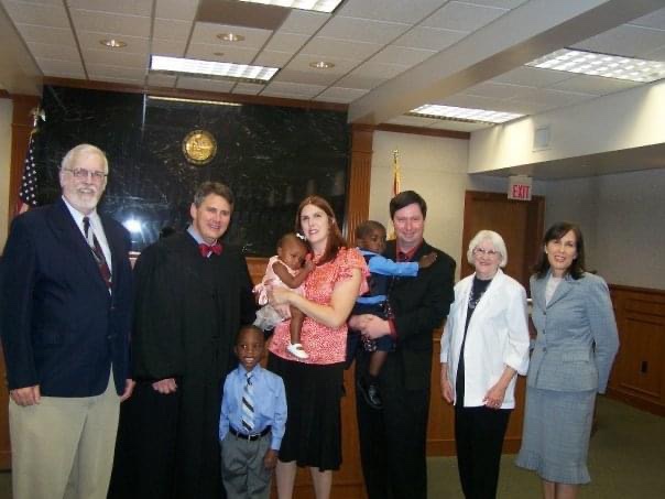 A happy family stands in a courtroom after an adoption ceremony, with a judge and several witnesses. The family includes two children held by adults, showcasing the joyful moment of finalizing their adoption.