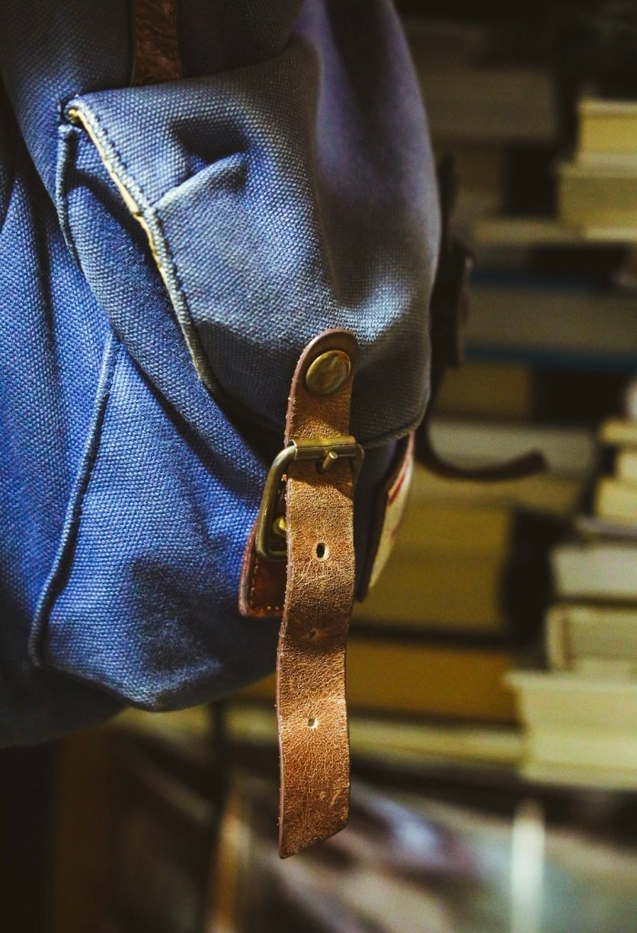Close-up of a blue backpack with a brown leather strap, set against a background of stacked books.