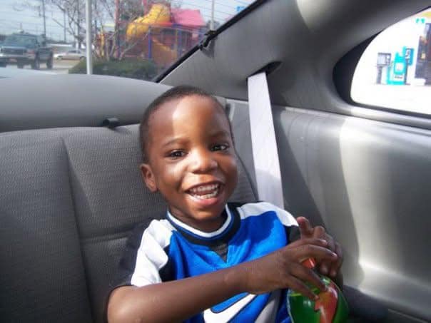 A smiling young boy sitting in the backseat of a car, holding a drink, with a playful expression on his face.