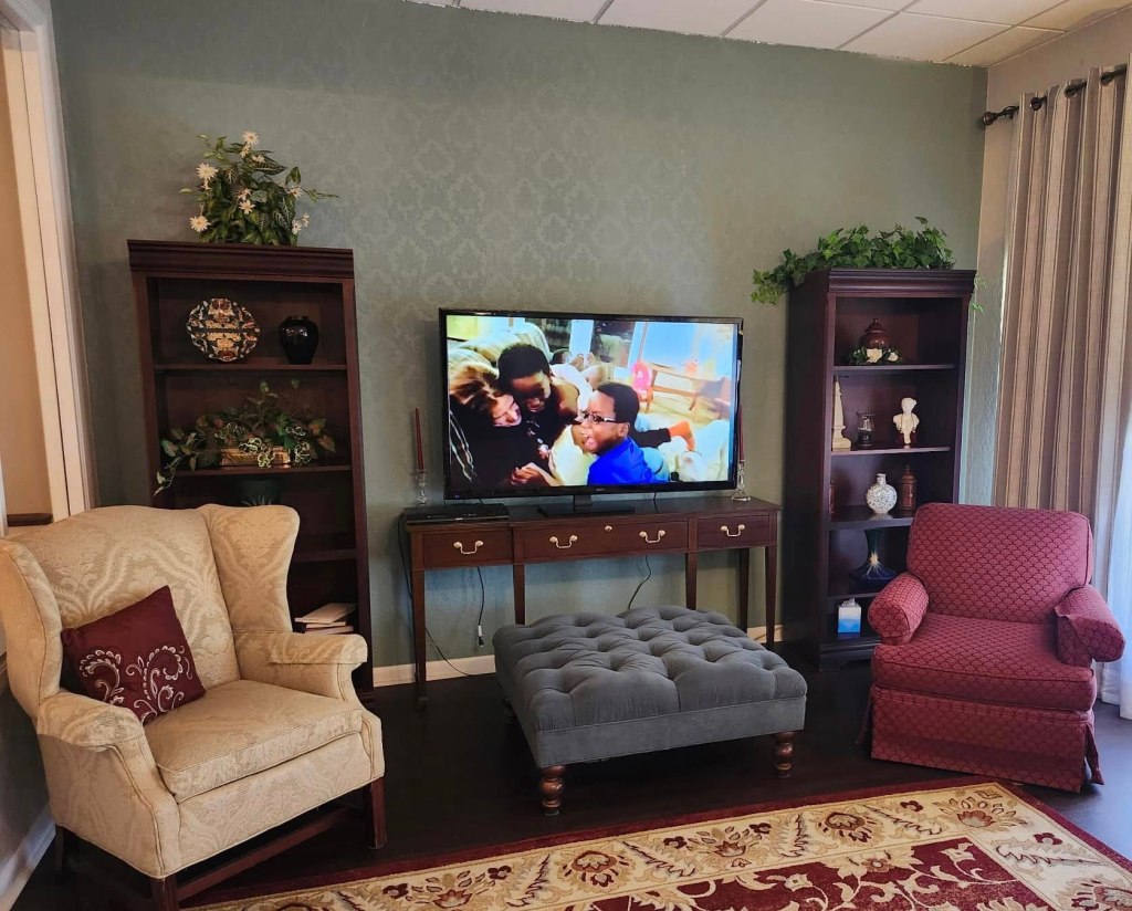 A cozy living room with two armchairs, a tufted ottoman, and a television displaying a family scene. Decorative shelving is visible, adorned with plants and ornaments.