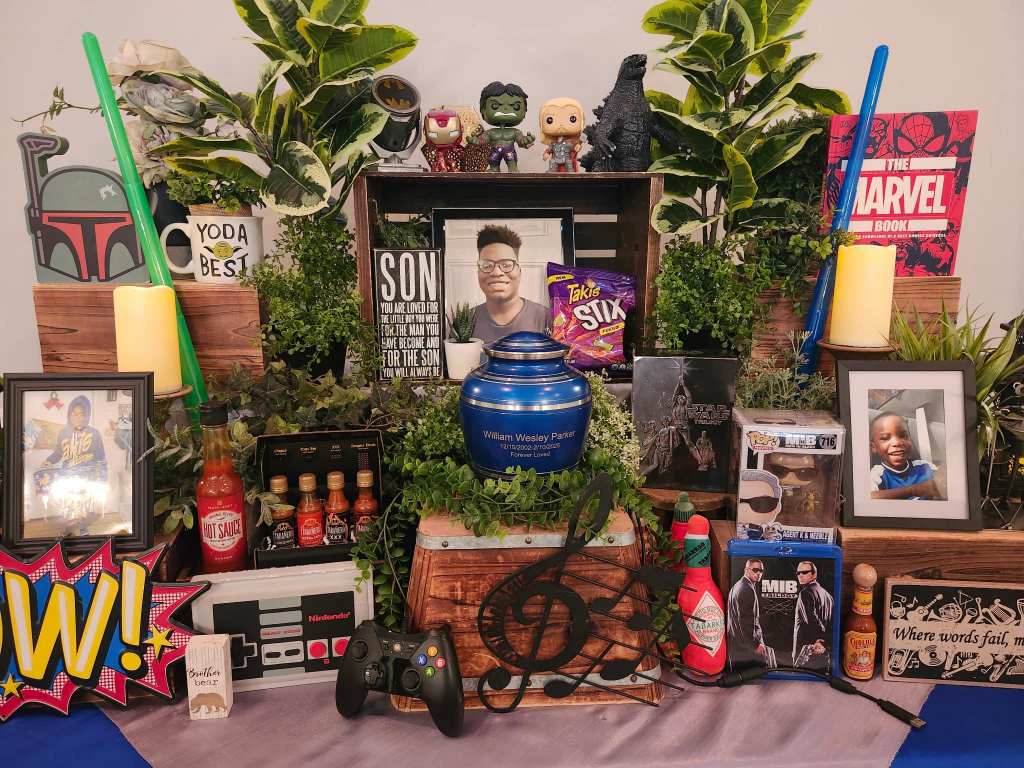 A memorial display featuring an urn, framed photos, candles, snacks, and various personal items honoring a loved one.