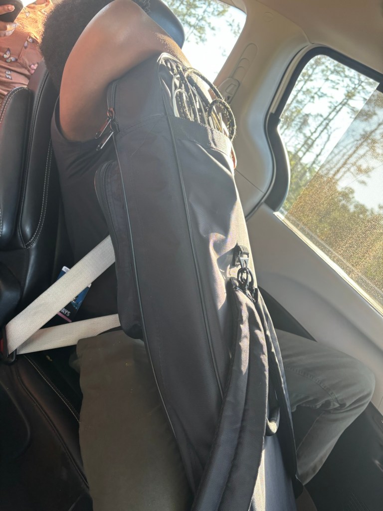 A person seated in a car, leaning against a black gig bag, with musical equipment visible inside.