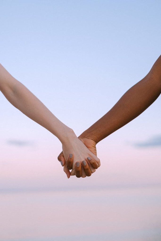 Two hands, one with light skin and the other with dark skin, interlocked against a pastel sky, symbolizing support and connection.