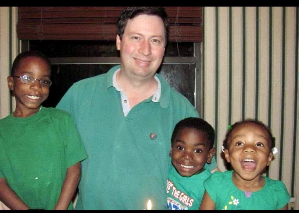 A man stands with three children, all smiling, in a cozy indoor setting. They are wearing green clothing, with two boys on either side of the man and a girl in the front, who is joyfully laughing. A candle is visible on the table, suggesting a celebration.