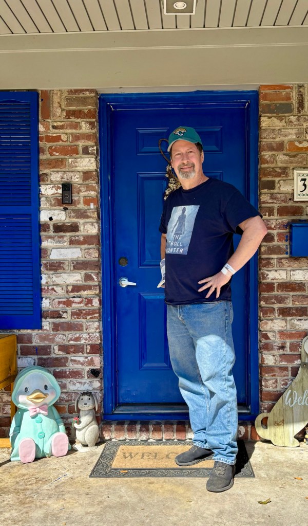 A man wearing a blue baseball cap and a dark shirt stands at a vibrant blue door, displaying a post-surgery wristband, with decorative garden statues on either side.