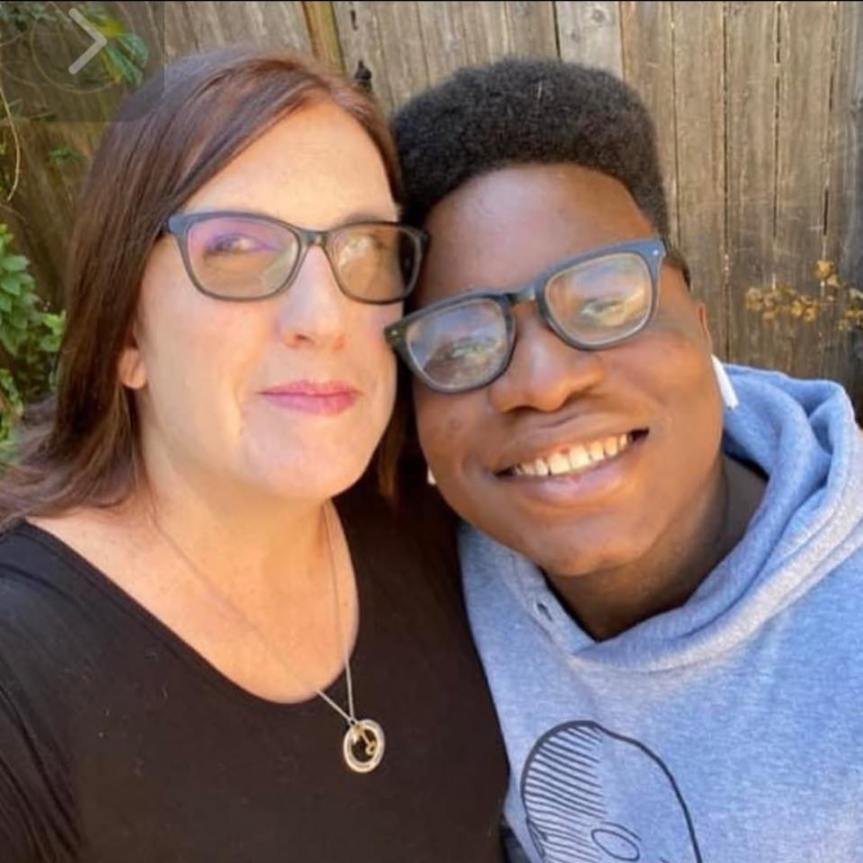 A close-up selfie of a woman with long brown hair wearing glasses and a black top, smiling alongside a young man with short black hair, also wearing glasses and a gray hoodie, both appearing joyful in front of a wooden fence.